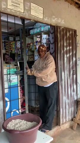 A woman wearing a brown hooded sweatshirt and black pants stands in the doorway of a small shop. She is smiling and gesturing with her hands. In front of the shop, a large brown plastic tub filled with what appears to be dried beans or grains sits on a white table. The shop has a metal gate that is partially open, revealing shelves stocked with various products. There are signs posted on the wall above the doorway. The shop appears to be made of corrugated metal.