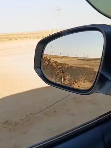 THE VIDEO SHOWS A REARVIEW MIRROR REFLECTING A DIRT ROAD IN A DESERT LANDSCAPE WITH POWER LINES IN THE BACKGROUND. THE MIRROR SHOWS A HOLE OR DITCH IN THE GROUND, WITH PILES OF DIRT NEARBY. THE CAMERA APPEARS TO BE INSIDE A MOVING VEHICLE.