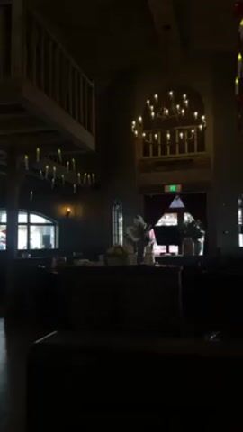 The video shows a dimly lit indoor space. A chandelier hangs from the ceiling, illuminating the space. There's a wooden balcony-like structure to the left with faux candles suspended beneath it. In the background, there is an archway draped with a red curtain, possibly leading to another area. Tables are visible, hinting at a restaurant or dining setting. People can be seen indistinctly through the window. The lighting creates a somber and mysterious ambiance.