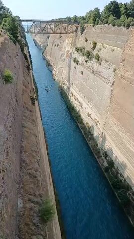 A drone shot captures a wide, deep canal with sheer, high cliffs on either side. The water in the canal is a vivid blue. A small sailboat is visible in the center of the canal, moving away from the camera. In the distance, a metal bridge spans across the canal. The sun shines brightly, casting shadows on the canyon walls. The vegetation on top of the cliffs is green and sparse.