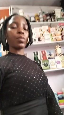 A young Black woman with braided hair is standing in front of shelves filled with beauty products. She is wearing a sheer black top with a diamond pattern. The shelves behind her are stocked with various items, including perfumes, lotions, and soaps. The lighting is bright, and the overall environment appears to be a retail store. The woman makes a few facial expressions and gestures throughout the short video.