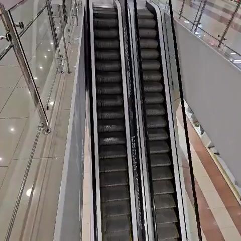 A high-angle shot shows two escalators in a shopping mall. The escalators have dark grey steps with black sides. The mall appears to be multi-leveled, with glass railings and shops visible on the upper level. The floor is tiled in a light color. The scene is well-lit, likely from overhead lighting in the mall. No people are visible on the escalators or in the immediate vicinity.