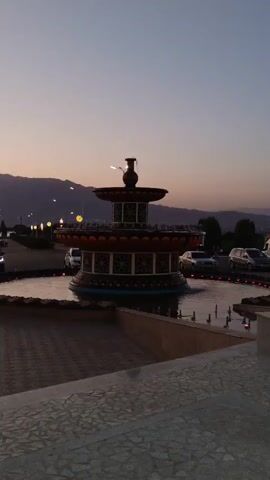 The video shows a dusk scene with a fountain in the foreground. The sky is a soft gradient of orange and purple, with mountains visible in the distance. Streetlights are on, illuminating a paved area with several cars parked and driving. The fountain has multiple tiers and is lit with colorful lights. The overall atmosphere is peaceful and serene.