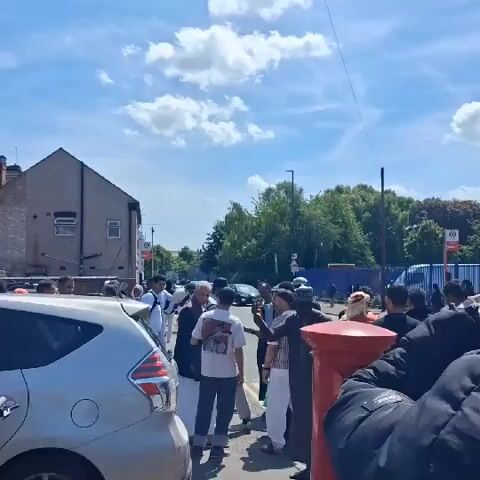 Here is a description of the video. 

The video shows a group of men gathered outside on a sunny day. Some of the men are wearing traditional Muslim clothing, including kufis or taqiyahs (skullcaps). They are standing near a street with parked cars, a red mailbox, and a brick building that appears to be a tire shop called "Tyres Plus". The sky is blue with scattered white clouds. People are walking around and talking. Some of the men look at the camera.