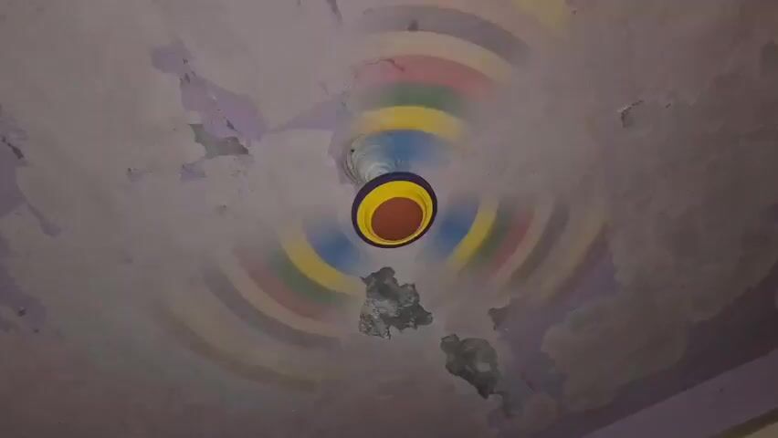 A ceiling fan spins rapidly against a textured, purple-painted ceiling. The fan has a white central housing and a blue, yellow, and orange decorative element. The ceiling itself shows signs of wear and tear, with patches of peeling paint and what appear to be water stains or damage. Concentric circles of various colors, including red, pink, yellow, green, and blue, are painted on the ceiling, radiating outwards from the fan, creating a visual effect that interacts with the fan's motion. The overall impression is one of a slightly dilapidated but colorful interior space.