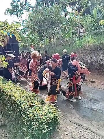 This is a medium-shot video of an outdoor performance. In the foreground, a person in a traditional costume, with a horse head mask and a striped shirt, is dancing. Behind them, other performers in similar costumes are also dancing and interacting. Some are holding props, possibly musical instruments or traditional weapons. In the background, there are trees, a speaker system, and a small group of spectators. The ground is wet, suggesting it might have rained recently. The atmosphere appears festive, with people actively engaged in the performance. The video is shot from a slightly elevated angle, looking down at the scene.