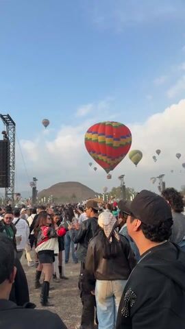 A crowd of people is gathered at an outdoor festival with multiple hot air balloons in the sky. The balloons are various colors and patterns, including one shaped like a smiley face. In the background, there appears to be a pyramid-like structure on a hill. There are speakers on scaffolding. Many people are looking up at the balloons.