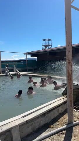 The video shows an outdoor pool area on a bright, sunny day. Several people, primarily adults, are immersed in the light-colored water of the pool. In the background, a building with a brown roof and a multi-level metal tower or platform is visible against a clear blue sky.

A prominent feature is a powerful stream of water spraying into the pool from the right side, creating a significant splash and mist. Several individuals are gathered near this spraying water, some seemingly enjoying the force of the spray, while others are farther away in the pool. The pool's edge appears to be made of concrete or stone. A wooden structure or platform is situated along the right side of the pool, adjacent to the water spray. A rusty metal pole stands in the immediate foreground on the right, partially obscuring the view. The scene suggests a relaxed atmosphere, with people enjoying swimming or bathing in what appears to be a thermal pool, given the vigorous water movement and the general context of such facilities in Zhemchug, Russia.