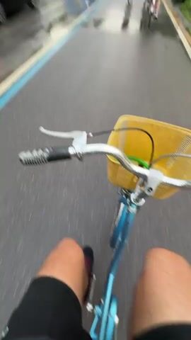 The video shows a first-person perspective of someone riding a bicycle on a wet road. The bicycle has a yellow basket on the front and the rider is wearing black shorts and red shoes. Other cyclists are also on the road, along with cars and a bus. The background features lush green scenery with unusual mountain formations under an overcast sky. There are roadside signs in Asian characters, and some small businesses visible on the side of the road. The road has a blue bike lane. The cyclist is moving at a moderate speed.