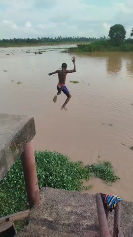 The video captures a person jumping into a murky brown river from a concrete structure. The subject, a young person wearing dark shorts, is initially seen standing at the edge with arms outstretched before leaping into the water. A significant splash erupts upon impact, followed by ripples spreading across the surface. The person then appears to be swimming or treading water, creating further disturbances in the water around them.

The scene is set in a natural environment with lush green vegetation growing along the riverbank and floating on the water's surface. In the background, a treeline with palm trees is visible, suggesting a tropical or subtropical climate. The sky appears overcast, indicating a cloudy day, possibly after a period of rain or before an approaching weather system. The overall impression is one of a spontaneous and adventurous act within a serene, rural landscape. There is no visible text in the video.