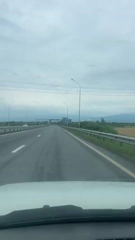 This media file captures a point-of-view shot from inside a vehicle traveling on a highway. The scene is a multi-lane road with a metal guardrail separating the lanes from the right shoulder. Several streetlights are visible along the highway, indicating it's likely a well-maintained route. On the right side of the road, there is a blue road sign with white text in Cyrillic. The sign indicates directions to "ВЛАДИКАВКАЗ" (Vladikavkaz) and "ВЕРХНИЙ ЛАРС" (Verkhny Lars), with "АРХОНСКАЯ" (Arkhonskaya) and "АРДОН" (Ardon) listed below, along with distances. The landscape outside the highway features greenery, including trees and fields, and in the distance, mountains are visible under a cloudy sky. The weather appears overcast, suggesting it might be a cloudy day. The time of day is not explicitly stated but the lighting suggests it is daytime. There are other vehicles on the road, including a white van in the left lane and other cars further ahead. The overall mood is one of travel and transit. The focus is on the road ahead and the directional information provided by the road sign, which places the location in proximity to Vladikavkaz, Verkhny Lars, Arkhonskaya, and Ardon in Russia.