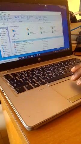 A single hand operates the touchpad of a silver HP laptop. The laptop rests on a wooden surface. The screen displays the File Explorer window in Windows, showing folders like "Downloads" and "Desktop," and "Recent files" including documents with titles such as "LONDON PRELIMINARY PADS" and "Notifications of change in Job Title." The hand is dark-skinned and its fingers are actively moving on the touchpad. The laptop's keyboard is visible, and other objects, including what appears to be another computer component, are visible in the blurred background.