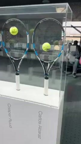 The video shows a display case containing tennis rackets and tennis balls, identified with the names of tennis players. The rackets appear to be the same model, predominantly black with blue accents, each with a tennis ball resting inside the racket head. The names associated with the rackets, from left to right, are "Stan Wawrinka," "Felix Auger-Aliassime," "Holger Rune," "Casper Ruud," and "Carlos Alcaraz." The case is well-lit, and the rackets are arranged in a row.  A child in pink clothing is seen briefly in the background, walking past the display.  The display case is white, and the names are printed along the base. The floor is gray.  The area appears to be a retail space or exhibit.