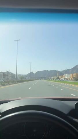 The video shows a driver's point of view while driving on a road with a speed limit of 80 km/h. The sky is clear and the sun is shining. There are buildings and mountains in the background. The road is well-maintained with lane markings. There are street lights lining the road. The dashboard and steering wheel of the car are visible.