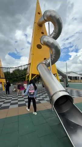 The video shows an outdoor playground with several large, yellow, cone-shaped structures resembling cheese wedges, complete with circular holes. Two of these structures are prominent in the foreground and midground. A winding, silver, metal slide extends from one of the cheese structures into the lower foreground of the frame. A net bridge connects two of the cheese structures in the midground. Several people, including children and adults, are present in the playground area. One person in the foreground is wearing a pink hooded outfit. The ground surface is a patterned mat with green, brown, and black and white checkered sections. The sky above is cloudy with some patches of blue.