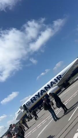 The video shows a Ryanair airplane on an tarmac under a bright blue sky with white clouds. Several people are walking towards the airplane, which appears to be boarding. The airplane's doors are open, and stairs are visible. Some people are carrying luggage. The tarmac is paved with white lines. The overall scene suggests a typical airport environment during daylight hours.