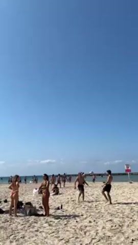 The scene takes place on a beach with light-colored sand and a clear blue sky. Several people are scattered across the beach, some standing, some sitting, and some walking. In the foreground, there are two young women in bathing suits standing near a bag. Two young men are playing with a ball in the middle ground. In the background, there are more people, and what appears to be a rocky structure. Overall, the scene depicts a sunny day at a populated beach.
