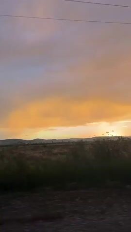 A vertical video shows a wide shot of a rural landscape at sunset. The sky is dominated by vibrant orange and yellow clouds, with hints of purple and pink. In the distance, silhouetted hills and trees are visible. A field of dry grass or crops stretches across the middle ground, appearing blurred due to motion. In the foreground, there is a dark, indistinct area that suggests the viewer is in a moving vehicle. A telephone pole with wires is positioned in the right-center of the frame, further emphasizing the rural setting. The overall atmosphere is serene and picturesque.
