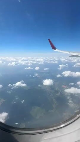 The video shows a view from an airplane window. A wing of the airplane with a red tip is visible in the upper right corner. Below the wing, a vast expanse of clouds stretches out, with patches of green land visible through gaps in the clouds. The sky is a clear blue. The video pans slightly, offering a changing perspective of the landscape and the clouds. The interior of the airplane, specifically the dark surrounding of the window frame, is also visible, casting a shadow on the window.