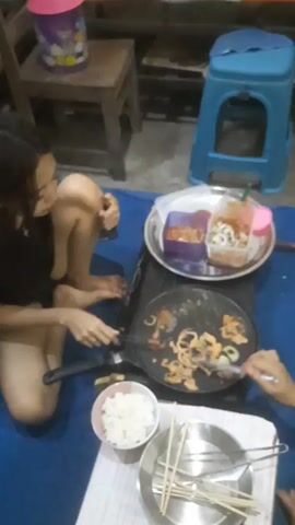 Two people are on the floor, preparing food. A person on the left, wearing a black shirt, is using tongs to stir food in a pan on a portable burner. A bowl of white rice is to the left of the pan. A metal plate with chopsticks and utensils is in front of the pan. To the right of the pan is a round tray holding two containers of food. The background shows wooden furniture and a blue plastic stool. The scene appears to be indoors.