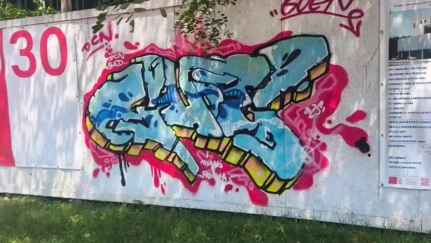 A wide shot of a graffiti-covered temporary wall shows a large stylized graffiti piece in shades of blue and yellow with pink spray paint drips and outlines. To the left of the graffiti is a large red number "30" on a white background, and to the right, a white panel with text and a diagram. The wall is outdoors, with green grass and trees visible in the foreground and background. The graffiti appears to be on a construction hoarding.