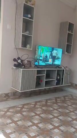 The video shows a living room or entertainment area with a tiled floor and a TV mounted on a wall-mounted cabinet. The floor has a pattern of brown, tan, and white tiles arranged in a geometric design. On the wall, there is a grey-colored wall-mounted TV cabinet with shelves and closed storage. A television is on the cabinet, displaying a scene from a show or movie. There are two tall, narrow shelves on either side of the TV, also mounted on the wall and matching the cabinet's grey color. A few decorative items sit on the shelves of the cabinet, and there's a computer tower visible inside one of the cabinet sections. The walls are a light neutral color. Electrical outlets and wires are visible on the wall. The lighting appears to be a combination of natural and artificial.