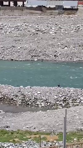 A river with turquoise water flows between rocky banks. Several people are visible swimming in the river, with one person standing on the bank. Buildings and vehicles are present in the background.