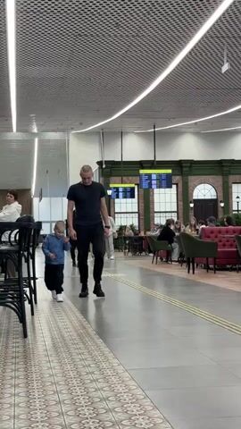 A man and a young boy are seen walking and running playfully through a spacious, modern indoor environment, likely a transport hub or large commercial area in Новая Казмаска, Russia. The man, dressed in a black t-shirt, black pants, and black shoes, walks a short distance behind the boy. The boy, wearing a blue hooded top, black pants, and white sneakers, initially walks, then turns to look at the man with a wide smile before breaking into a run, his arms swinging. The man responds with a smile.

The setting features a high ceiling with slatted acoustic panels and long, linear lights. The floor is tiled with a mix of plain gray and patterned sections. To the right, there is a large seating area, resembling a cafe or restaurant, furnished with tables, chairs, and upholstered booths in red and dark green. A long counter with black high stools and decorative hanging lamps is also visible. In the background, large digital screens display information, with visible text including "Рейсы" (Flights) and "ВРЕМЯ" (Time). Another wall segment shows "НЕДЕЛЬНЫЙ ОБОРОТ" (Weekly turnover), and a partial sign reads "KUK...". Other objects include a white baggage cart and stacks of bottled water. The overall impression is one of a bright, well-lit indoor space, and the interaction between the man and boy suggests a happy, casual moment.