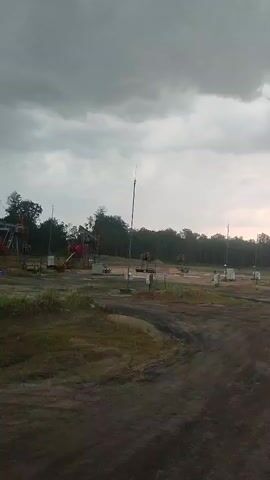 The video shows a landscape with several oil pumpjacks. The sky is overcast and appears to be cloudy or stormy. The ground is dirt and grass, with some areas that look like they have been cleared or disturbed. There are trees in the background. The oil pumpjacks are spaced out across the area, and some appear to be in operation. The overall impression is an industrial setting within a natural environment.