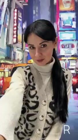 A medium shot shows a woman with black hair in a ponytail, wearing a cream-colored turtleneck and a beige sweater vest with a black animal print, looking at the camera. The background appears to be a colorful painting of a city scene with a yellow taxi cab. The lighting is bright and even.