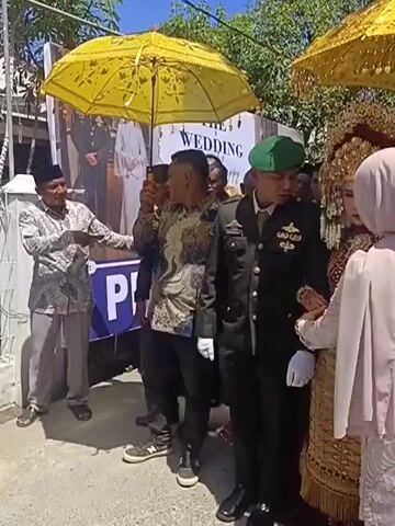 The video shows a wedding procession outdoors. In the foreground, a man in a military uniform holds the hand of a woman adorned in traditional wedding attire, including an elaborate headdress and ornate clothing. To their left, another man, possibly a relative or escort, holds a yellow umbrella over them. Several other individuals are visible in the background, some also holding yellow umbrellas. A banner with the word "WEDDING" is partially visible behind the procession. The environment appears to be a street with buildings and trees under a clear sky. The overall atmosphere seems to be celebratory.