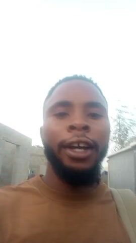 A man with dark skin, a beard, and a tan t-shirt is speaking to the camera, smiling and pointing with his index finger. He states "off to work, all time baby" and then "all time baby" again while grimacing. The background shows cinderblock construction and a hazy sky. A strap for a bag hangs over his right shoulder.