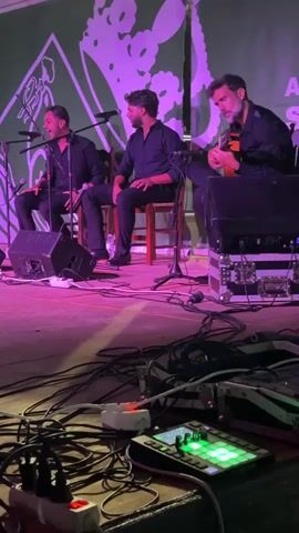 The video shows three men on a stage performing. The man on the left is singing into a microphone and playing a percussion instrument. The man in the middle is also singing into a microphone and is seated on a chair. The man on the right is playing a guitar and is seated on a chair. The stage is lit with purple lighting. In the foreground, there are several audio cables and a piece of audio equipment with illuminated green buttons. The background features a green curtain with a white graphic design.