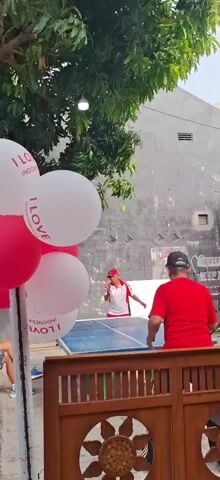 The video shows two men playing table tennis outdoors. The man on the left is wearing a red and white shirt and a red cap. The man on the right is wearing a red shirt and a black cap. They are both facing each other across the table. There are balloons in the foreground with the words "I LOVE INDONESIA" printed on them. The background consists of a gray wall with some graffiti and a tree. The overall setting appears to be a casual outdoor event.