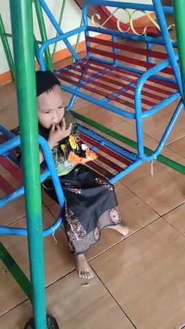 A child is sitting on a swing set. The child is wearing a black cap, a green and yellow patterned shirt, and a black sarong. The child is looking towards the camera and has their hand near their mouth. The swing set is blue and red with green poles. The ground is tiled with tan tiles. There is a wall behind the swing set.