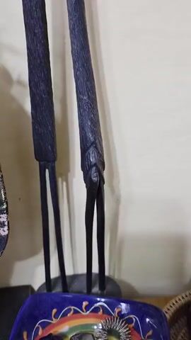 A close-up shot focuses on two dark, stylized wooden figurines. The first figurine, on the left, depicts a person with a hand raised to their mouth, appearing to be in thought or conversation. The second figurine, to the right, is more abstract, with a slender body and no discernible facial features. The figures are positioned in front of a plain, light-colored wall. The lighting is soft, casting subtle shadows. The overall impression is one of a still-life arrangement, possibly on a shelf or display.