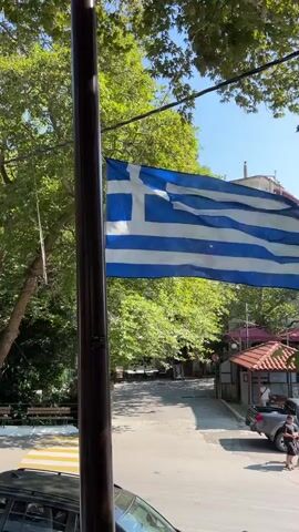 A Greek flag waves in the wind against a backdrop of lush green trees and a clear blue sky. A dark pole stands to the left of the flag, obscuring part of it from view. In the background, a street is visible with a pedestrian crossing and a few vehicles, including a gray truck. There are also glimpses of buildings and covered seating areas. The overall scene appears to be outdoors on a sunny day, possibly in a town or village setting.