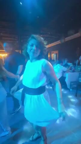 A woman with brown, curly hair and a white dress dances on a lit dance floor in a dimly lit venue. The woman is wearing a black belt around her waist and black high-heeled shoes. She is smiling and appears to be enjoying herself. In the background, other people are seated at tables and standing around the room. The venue appears to be a restaurant or club, with tables, chairs, and decorative elements visible. The lighting is dynamic, with spotlights and colored lights illuminating the dance floor and surrounding areas. The video captures the woman's movement from different angles as she dances.