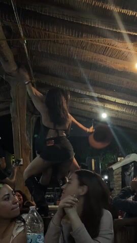The video shows two women dancing on what appears to be a stage or elevated surface, in a dimly lit environment. The women are facing away from the camera; one is wearing dark shorts, a top that exposes her back, and tall boots, while the other is wearing a cowboy hat. The women are embracing each other as they move. Several people are seated at tables, watching the women and recording them with their phones. A bottle of water is visible on a table in the foreground. The ceiling is made of straw or similar organic material. The scene appears to be taking place in a bar or outdoor venue.