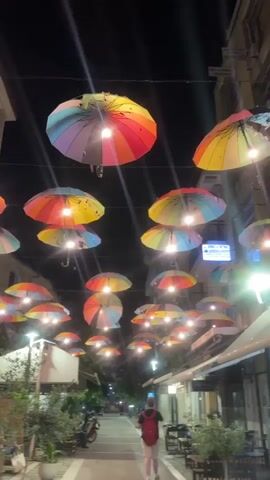 The video captures a night scene on a pedestrian street in Karditsa, Greece. Numerous brightly colored, rainbow-patterned umbrellas are suspended overhead, forming a decorative canopy. Many of these umbrellas have lights attached to their undersides, illuminating them and casting a warm glow onto the street below. Additional streetlights are visible along the sides of the street, further brightening the area.

The street is lined with buildings, presumably housing businesses, as evidenced by outdoor seating areas with tables and chairs visible on the right side. Potted plants and small trees are placed along the sidewalks, adding greenery to the urban environment. In the distance, a person wearing a red backpack walks away from the camera, down the illuminated path. A motorcycle or scooter is parked on the left side of the street.

Signs with Greek text are visible on the buildings, including "ΑΣΦΑΛΕΙΕΣ Α. ΔΡΟΥΓΚΑΣ" and "ΣΧΟΛΗ ΟΔΗΓΩΝ Α. ΧΡΗΣΤΟΥ," confirming the Greek location. Another sign on the right building displays "PAPACOULIS." The dark sky indicates it is nighttime, with the artificial lighting providing the main illumination for the vibrant streetscape. The overall atmosphere appears lively and inviting.