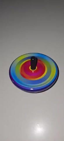 A close-up, high-angle shot shows a colorful spinning top on a white surface. The top has a black stem and a circular body adorned with concentric rings of red, orange, yellow, green, blue, and purple. The spinning motion blurs the colors into a rainbow effect. The background is a plain white surface, with slight variations in lighting that create soft shadows. The lighting is bright and appears to be coming from above and to the right. There are no people or other objects visible in the frame.