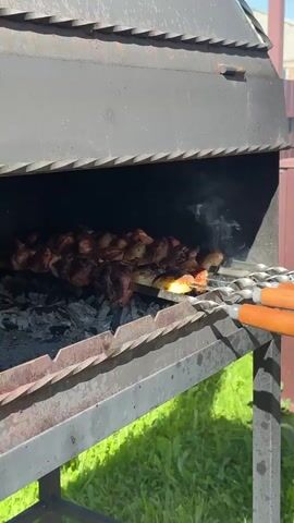 The video shows a close-up of a black metal barbecue grill with cooked meat on skewers inside. The skewers have wooden handles and are resting on the side of the grill. Below the skewers are hot coals, with some smoke rising. The grill is sitting on green grass, and a brown fence and a house can be seen in the background. It is a bright, sunny day.