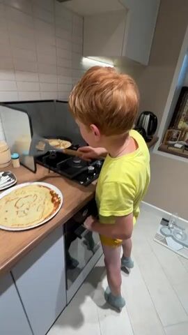 This media file captures a young boy with red hair, wearing a yellow t-shirt and yellow shorts, in a kitchen. He is standing in front of a stove with a frying pan, actively cooking. He is holding a spatula and appears to be flipping or moving a pancake in the pan. Next to the stove, on the kitchen counter, is a plate piled high with several cooked pancakes. The kitchen has white tiled backsplash, a wooden countertop, and a light-colored cabinet. In the background, a kettle and what looks like an icon are visible on a shelf. The scene appears to be indoors, likely in a home kitchen, given the domestic setting and the presence of cooking activities. The lighting suggests it could be daytime, as there is ample light coming from above and possibly a window to the right. The boy's focus on the cooking suggests he is engaged in the task, and there's a sense of domestic activity or perhaps a child learning to cook. There is no visible text in the media file. The location context provided is "Пычанки, Russia," which places the scene within a specific geographical region.