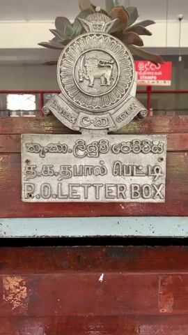 This media file shows a close-up view of a vintage wooden post box in Nuwara Eliya, Sri Lanka. The post box is a rich, dark wood with some signs of age and wear, including scratches and scuffs. At the top of the post box is a circular metal emblem featuring a lion with a sword, a common symbol in Sri Lanka, and the word "CEYLON" below it. Below the emblem, a metal plaque displays text in three different scripts: a Tamil script, another script that appears to be Sinhala, and "P.O. LETTER BOX" in English. Further down the post box, a rectangular label reads "INTERNATIONAL AIRMAIL & OVERSEAS". Another plaque at the very bottom, partially obscured, also contains text in Sinhala and English, likely indicating "LETTER BOX". The setting appears to be outdoors, possibly at a post office or a public area where mailboxes are located. There are no people visible in the frame. The lighting suggests it could be daytime, with natural light illuminating the post box. The overall impression is one of history and nostalgia, with the post box serving as a tangible link to past postal services in Sri Lanka.