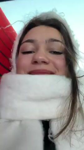 A young woman is recording a selfie video inside what appears to be a bus. She is wearing a white, furry hat with red decorations and a thick, white scarf. The woman smiles and poses slightly for the camera.  She later blows a kiss. The background includes other bus seats, a window, and a red surface.