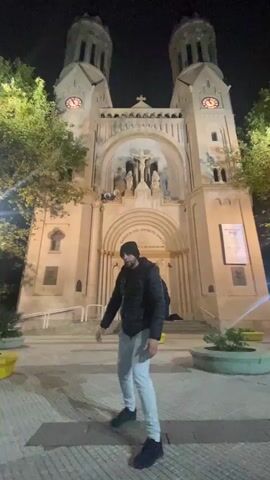 At night, a person with a black hat and a black jacket over a light gray trousers stands in front of the Basilica del Espiritu Santo. The Basilica is illuminated, revealing a facade with two tall towers, a central archway featuring religious statues and a cross at the top. The person moves their arms rhythmically, in what appears to be a dance movement. The surrounding environment includes trees and paved ground with planters.