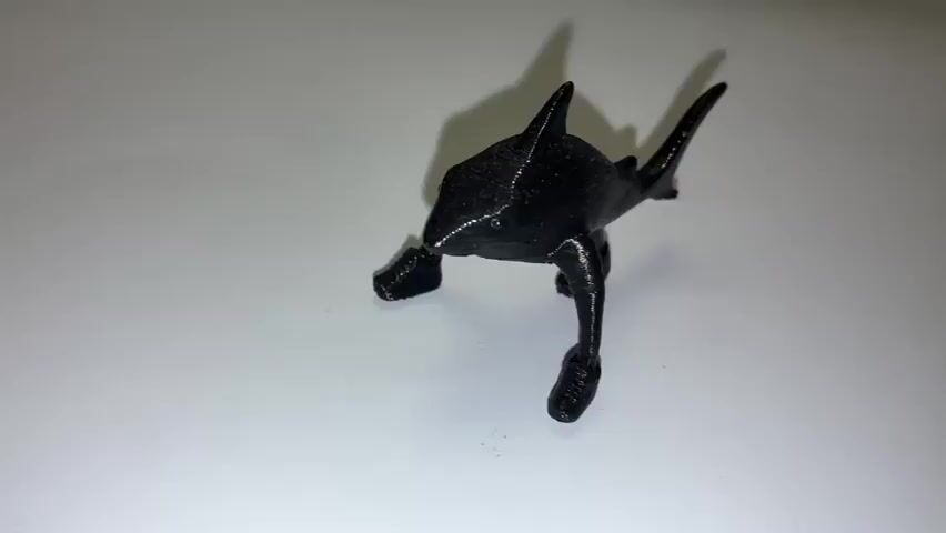 A black, 3D-printed toy resembling a shark with legs is positioned on a white surface. The toy is shown from a low-angle perspective, with its head facing the camera and its body slightly tilted. The background is a plain white surface, with a shadow cast by the toy. The lighting is even, highlighting the texture of the 3D print.