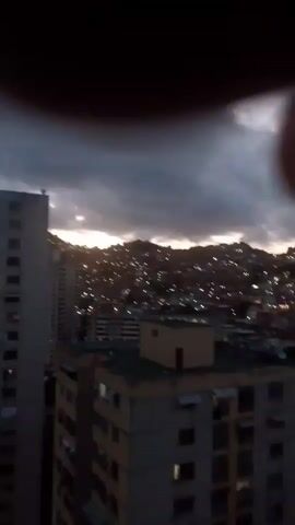 This is a short, low-resolution, handheld video clip, showing a city scene at dusk or dawn. The camera is tilted upwards, and the footage is shaky. In the foreground, there are parts of two buildings. The building closest to the viewer is mostly out of frame, showing only a corner of its side. The second building is a tall, multi-story apartment building, with visible windows and balconies. Its facade is a pale yellow color. On top of this building, there is a smaller, square structure with a flat roof, colored a light yellow or beige.

In the middle ground and background, a densely populated hillside is visible. Numerous small lights, appearing as streaks or blurs due to the low light and camera movement, illuminate the hillside, suggesting a favela or informal settlement. The lights are a mix of yellow and white. The sky above is overcast with dark, heavy clouds, but a sliver of lighter sky, possibly indicating the rising or setting sun, is visible on the left horizon behind the buildings. The overall impression is of an urban environment at twilight, possibly with a contrast between more established buildings and informal housing on the hills.