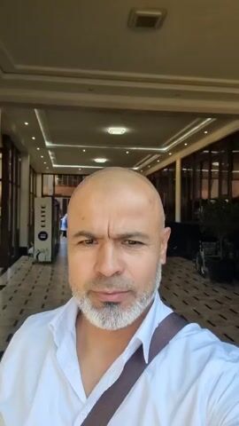A bald, middle-aged man with a gray beard wearing a white shirt is taking a selfie video. He is standing in front of a building with large glass doors and then moves to an outdoor location with trees and a street. He is carrying a brown strap over his shoulder. The video captures him from the chest up, focusing on his face as he turns his head slightly.