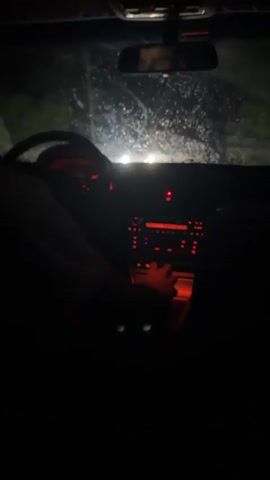 The video shows the interior of a car at night from the passenger's perspective, looking towards the dashboard and the driver. The driver's left hand is visible on the steering wheel. The dashboard lights are illuminated in red. A hand is operating the gear shifter. The windshield is visible with some light reflections, suggesting it is raining or wet. The rear-view mirror shows a partial reflection of a person's face. The environment outside the car is dark, with some faint lights visible in the distance. The camera is unsteady, indicating movement.